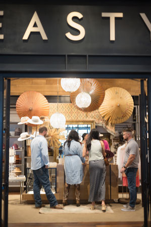 Shoppers at a trendy retail boutique counter in a mall, surrounded by decorative paper parasols, sparkling hanging chandeliers, a hat display and clothing racks.