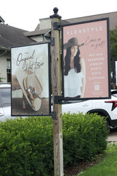 Outdoor double-sided retail sign mounted on a wooden post showing a smiling woman wearing a cowboy hat on one panel and a decorated Western hat on the other, set above green hedges and parked cars.