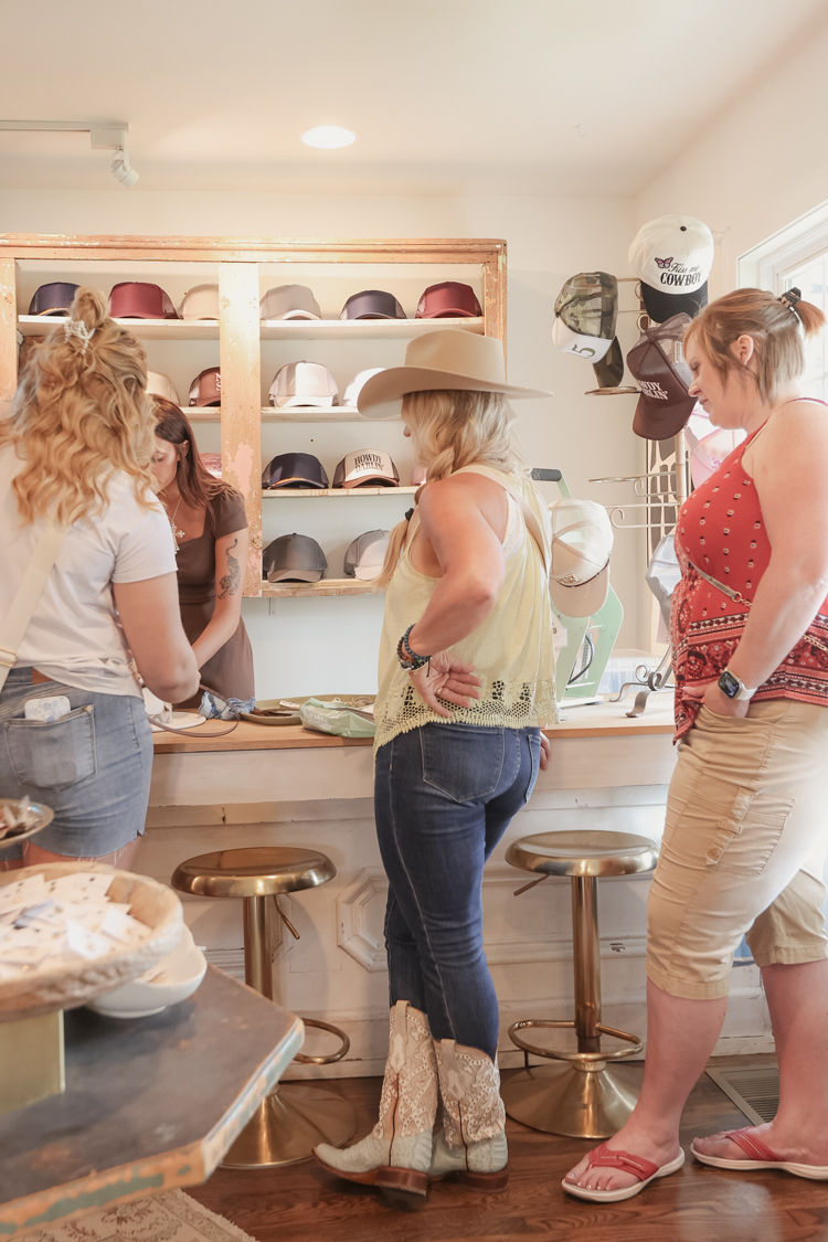 Three women browsing hats in a sunny boutique — shopper wearing a wide-brim cowboy hat and lace cowgirl boots at a counter with a wall display of caps.