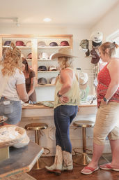 Three women browsing hats in a sunny boutique — shopper wearing a wide-brim cowboy hat and lace cowgirl boots at a counter with a wall display of caps.