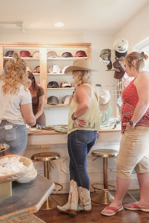 Three women browsing hats in a sunny boutique — shopper wearing a wide-brim cowboy hat and lace cowgirl boots at a counter with a wall display of caps.
