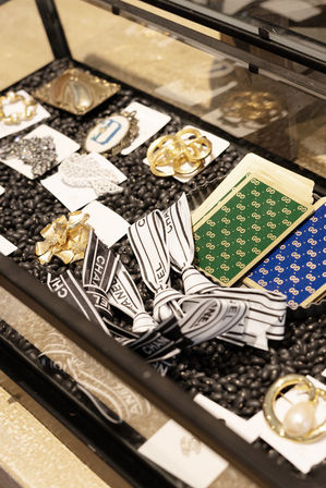 Boutique jewelry display case with assorted fashion accessories — gold and pearl brooches, decorative pins, patterned cards, and black-and-white ribbon bows on a bed of black beads.
