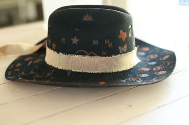 Black wide-brim felt western-style hat with a cream ribbon band and playful metallic copper stars, letters and shapes, resting on a sunlit white wooden table.