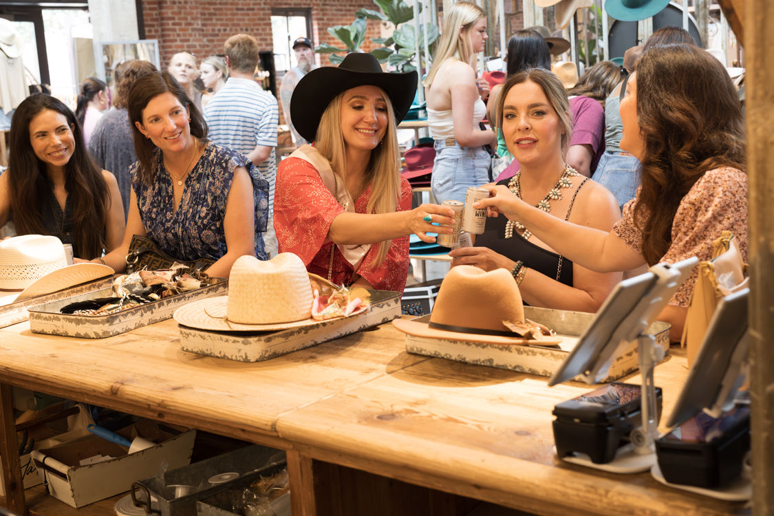 Group of smiling women at a wooden counter inside a brick-walled hat shop, trying on straw and felt hats—one wearing a black cowboy hat—clinking canned drinks amid displayed hats and busy boutique shoppers.