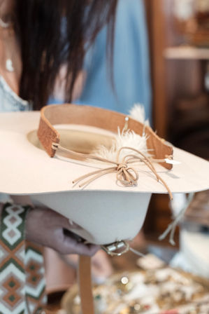 Light beige felt wide-brim hat with tan leather band, feather accent and tied suede cord held over a shop display — boho-western women's accessory.