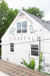 White gabled storefront with a painted hat and boot mural around a large upper window, white brick walls, green vines near utility meters, translucent corrugated awning and leafy trees in the background.