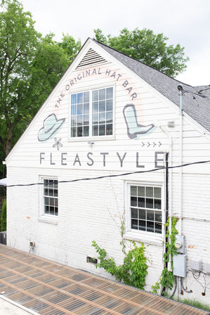 White gabled storefront with a painted hat and boot mural around a large upper window, white brick walls, green vines near utility meters, translucent corrugated awning and leafy trees in the background.