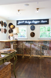 Bright cozy boutique interior with a wooden counter and patterned front, four brass bar stools, wall of hats and a neon sign reading "good things happen here" above two windows with leafy view.