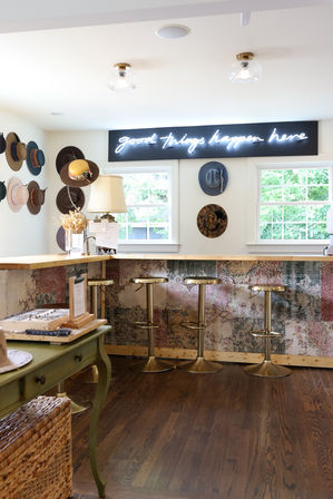 Bright cozy boutique interior with a wooden counter and patterned front, four brass bar stools, wall of hats and a neon sign reading "good things happen here" above two windows with leafy view.