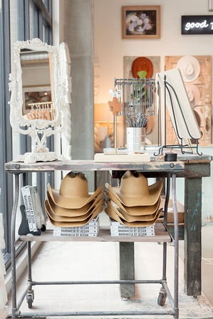 Bright home-decor boutique display with stacked straw cowboy hats on a rustic metal table, ornate white mirror and design books