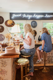 Two women seated at a wooden counter in a bright hat boutique, trying on and crafting wide-brim hats beneath a neon "good things happen here" sign.