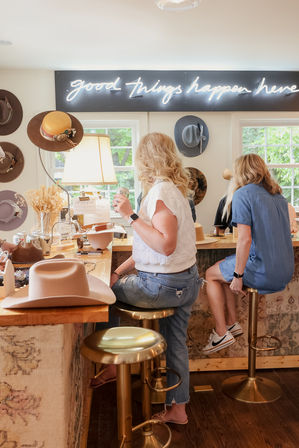 Two women seated at a wooden counter in a bright hat boutique, trying on and crafting wide-brim hats beneath a neon "good things happen here" sign.