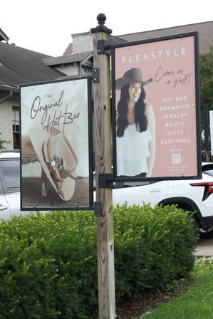 Outdoor double-sided sign on a wooden post advertising a hat boutique — one panel shows a smiling woman in a cowboy hat, the other a close-up of a decorated cowboy hat; suburban storefront, parked cars, and green shrubs in the background.