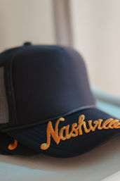 Black trucker cap with orange embroidered Nashville script on the curved brim, soft-lit close-up