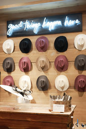 Boutique hat shop interior with a neon 'good things happen here' sign above a wooden wall displaying rows of colorful felt cowboy hats, plus a decorative wide-brim hat and hat brushes on a wooden counter.