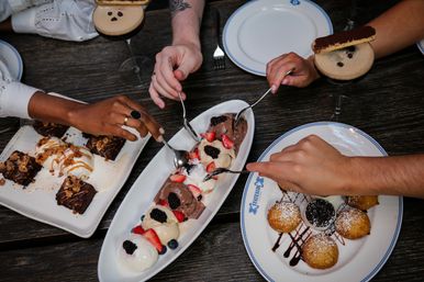 Several hands reaching with spoons over a dark wooden restaurant table filled with desserts — a long platter of chocolate and vanilla ice cream scoops topped with strawberries and blackberries, brownie bites with caramel and nuts, powdered-sugar fried dough balls with chocolate drizzle, and espresso martinis garnished with cookies.
