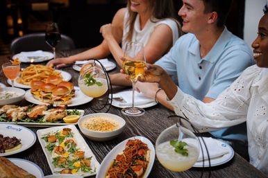 Group dining at a lively urban bistro: people toasting with white wine over a table of sliders, fries, scallops, grilled dishes and cocktails.