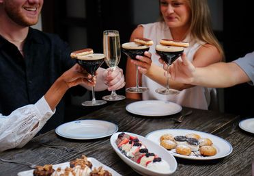 Four people clinking espresso martinis (topped with cookies) and a champagne flute above a wooden restaurant table filled with plated desserts — mini doughnuts, berry-topped cream and chocolate bites.