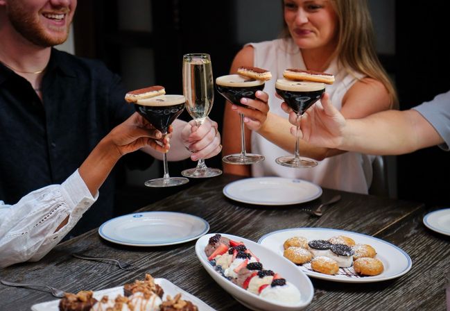 Four people clinking espresso martinis (topped with cookies) and a champagne flute above a wooden restaurant table filled with plated desserts — mini doughnuts, berry-topped cream and chocolate bites.