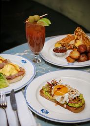 Colorful brunch spread: avocado toast with a sunny-side-up egg, eggs benedict, a breakfast burrito with roasted potatoes, and a Bloody Mary garnished with olives and lime on blue-rimmed plates.