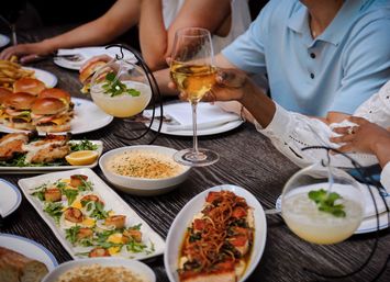 Group dining at a restaurant table featuring shared small plates and cocktails — sliders, seared scallops, baked gratin, roasted salmon, fries, hanging mint cocktails, and a raised glass of white wine.