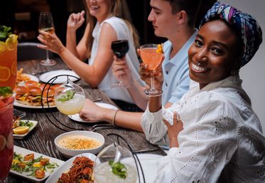 Group of friends at a restaurant table — smiling woman in a patterned headscarf toasting with a pink cocktail, surrounded by shared small plates, sliders and seafood.