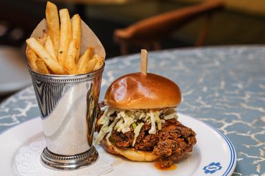 Crispy fried chicken sandwich with creamy coleslaw on a brioche bun served alongside golden French fries in a silver cup on a blue-patterned table.