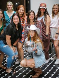 Smiling group of women at an indoor party venue on black-and-white geometric tile; one crouches in a white cowboy hat and boots sipping from a mirrored disco-ball punch bowl with a blue straw — playful friends' night scene.