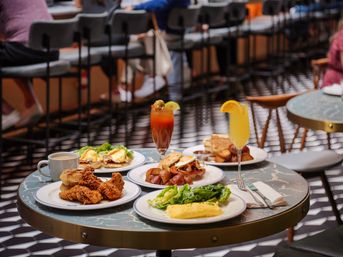 Colorful brunch spread at a busy downtown cafe — fried chicken, eggs Benedict, omelette with greens, roasted potatoes and toast, plus a Bloody Mary and mimosa on a marble-top table.