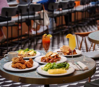 Colorful brunch spread at a busy downtown cafe — fried chicken, eggs Benedict, omelette with greens, roasted potatoes and toast, plus a Bloody Mary and mimosa on a marble-top table.