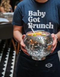 Person holding a disco ball mirrored punch bowl filled with pink cocktail, citrus wedges and a ladle, wearing a navy t-shirt that reads "Baby Got Brunch" in a lively brunch setting.