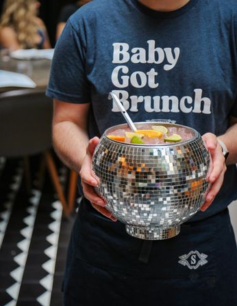 Person holding a disco ball mirrored punch bowl filled with pink cocktail, citrus wedges and a ladle, wearing a navy t-shirt that reads "Baby Got Brunch" in a lively brunch setting.