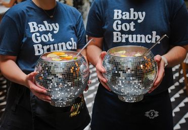 Two servers wearing "Baby Got Brunch" t-shirts hold mirrored disco‑ball punch bowls filled with iced cocktails and citrus garnishes at a lively brunch