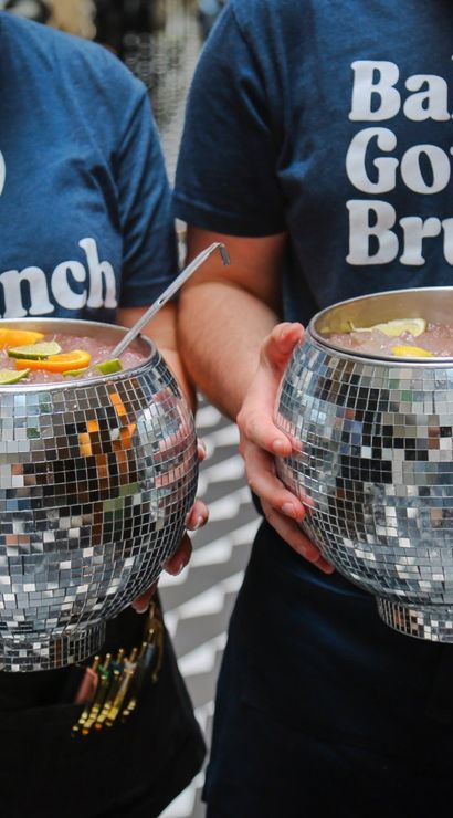 Two servers wearing "Baby Got Brunch" t-shirts hold mirrored disco‑ball punch bowls filled with iced cocktails and citrus garnishes at a lively brunch