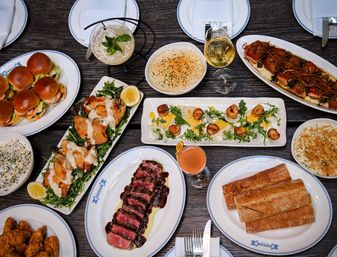 Overhead restaurant table spread featuring sliced steak with sauce, seared scallops on arugula, mini sliders, roasted fish with noodles, crusty bread, creamy sides, fried chicken, wine and cocktails on a dark wood table.