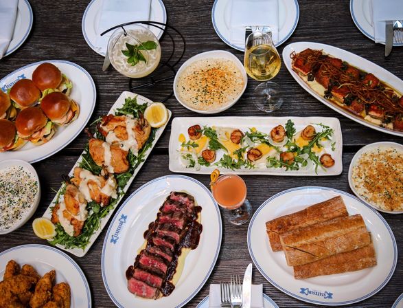 Overhead restaurant table spread featuring sliced steak with sauce, seared scallops on arugula, mini sliders, roasted fish with noodles, crusty bread, creamy sides, fried chicken, wine and cocktails on a dark wood table.