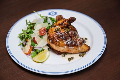 Grilled half chicken with caper cream sauce, served with arugula and cherry tomato salad topped with shaved Parmesan and a lemon wedge on a white rimmed plate