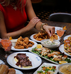 Diner in a red top dips a potato chip into herbed dip at a restaurant table surrounded by fried chicken, steak, sliders, salad and pink cocktails — casual shared appetizers and dinner scene.
