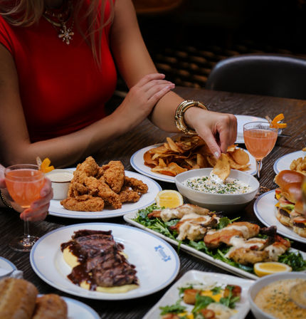 Diner in a red top dips a potato chip into herbed dip at a restaurant table surrounded by fried chicken, steak, sliders, salad and pink cocktails — casual shared appetizers and dinner scene.