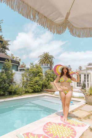 Person in a yellow floral bikini and oversized rainbow sun hat standing on a pineapple beach towel beside a small suburban backyard pool, fringed umbrella overhead, palm tree and blue sky in the background.