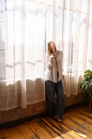Sunlit industrial loft with tall windows and sheer curtains; barefoot woman in an oversized white shirt and jeans strikes a relaxed pose beside a brick wall and potted monstera on a warm wooden floor.
