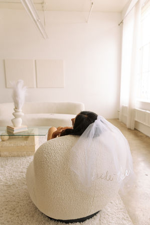 Bride-to-be lounging on a cream bouclé chair in a bright minimalist living room, wearing a short white veil, with a glass coffee table, vase and pampas grass nearby.