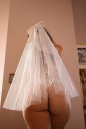 Cheeky bridal boudoir shot — rear view of an adult wearing a sheer white veil and tulle over bare hips against a soft pink wall.