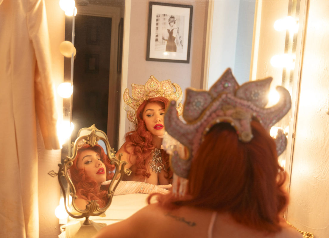 Glamorous red-haired performer adjusting a jeweled crown at a lighted vanity mirror in a backstage dressing room, multiple reflections visible.