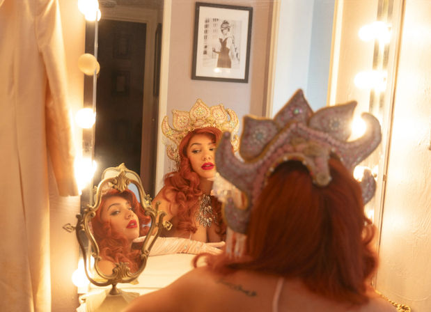Glamorous red-haired performer adjusting a jeweled crown at a lighted vanity mirror in a backstage dressing room, multiple reflections visible.