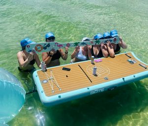 Six friends in bikinis and matching blue caps stand in clear shallow green water around a teal-edged inflatable floating platform, holding a long decorated plank with flamingo, palm and sea-themed stickers while enjoying canned drinks.