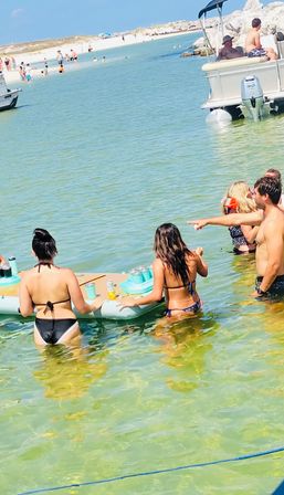 Group in swimsuits gathered around an inflatable floating drink table in clear shallow coastal water with a pontoon boat and sandy beach in the background on a sunny beach day.