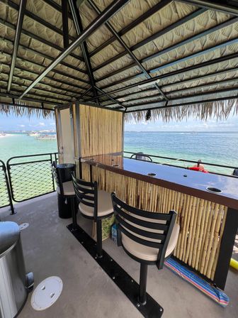 Overwater tiki bar deck with thatched roof, bamboo counter and cushioned bar stools overlooking turquoise ocean and boats in the distance