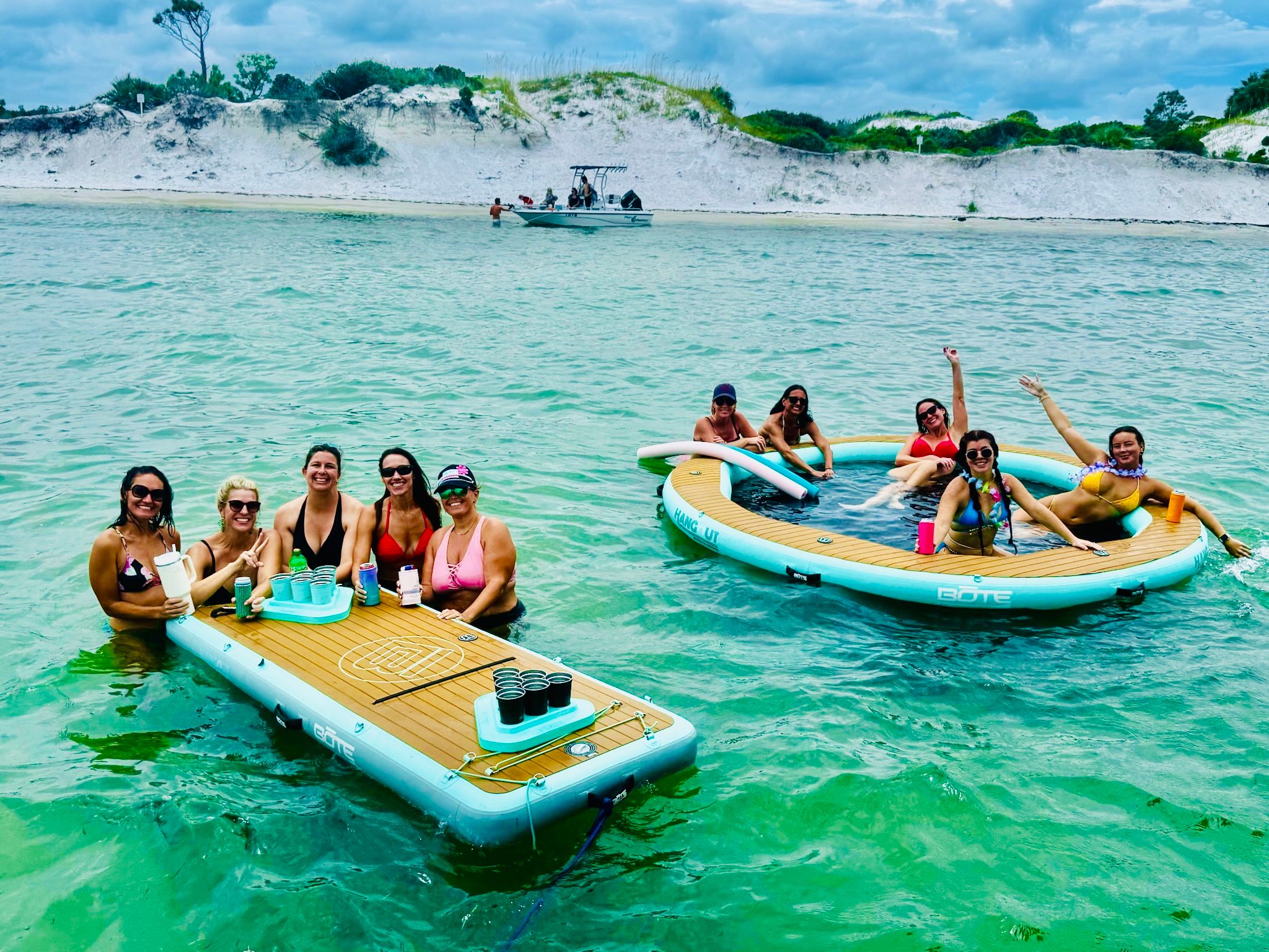Group of friends laughing and holding drinks on inflatable floating platforms in clear turquoise coastal water near white sand dunes and a small boat offshore
