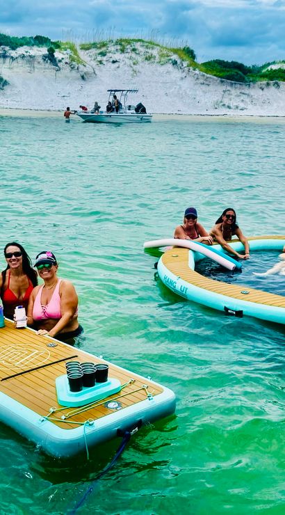 Group of friends laughing and holding drinks on inflatable floating platforms in clear turquoise coastal water near white sand dunes and a small boat offshore